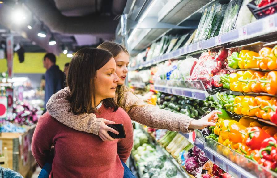 A young girl riding on her mother's back reaches for a pepper in the grocery store, holding a cell phone in her other hand