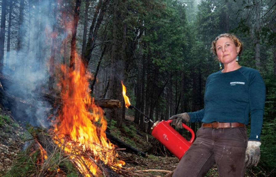 A woman stands next to a controlled fire she has lit in a forest
