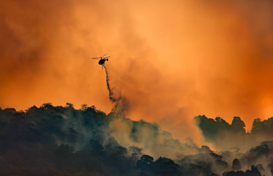 A helicopter dropping fire retardant on a burning hillside, flying through an orange, smoke-filled sky