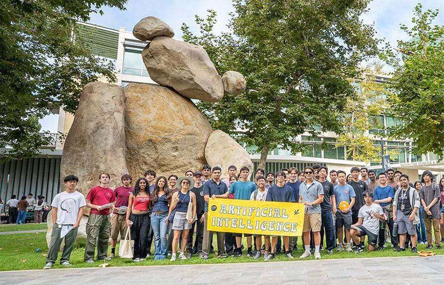 Dozens of students stand in front of a large stone sculpture in a courtyard, holding a banner reading ARTIFICIAL INTELLIGENCE