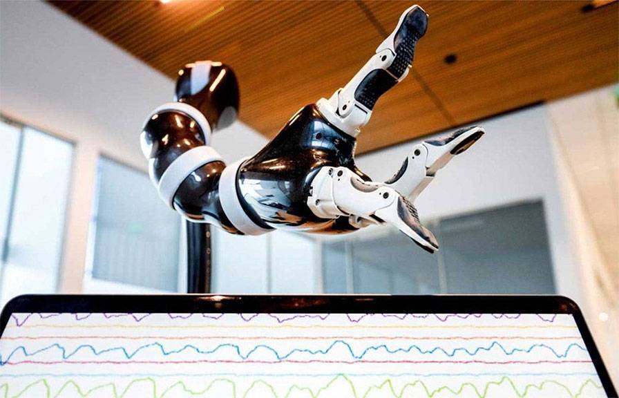 A robotic prosthetic hand with articulated black-and-white fingers hovers above a laptop screen displaying multicolored brainwave-style graphs in a modern indoor setting.