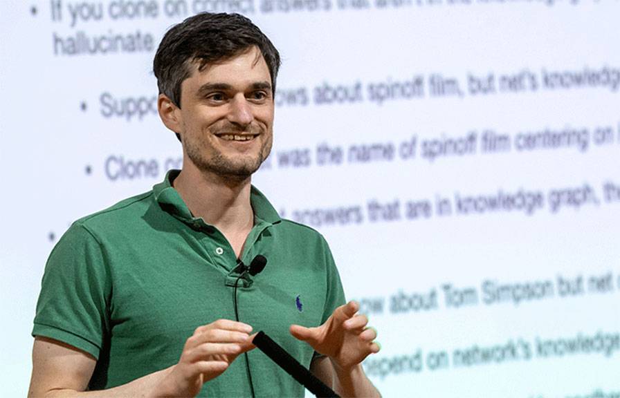 John Schulman speaks animatedly in front of a projected powerpoint
