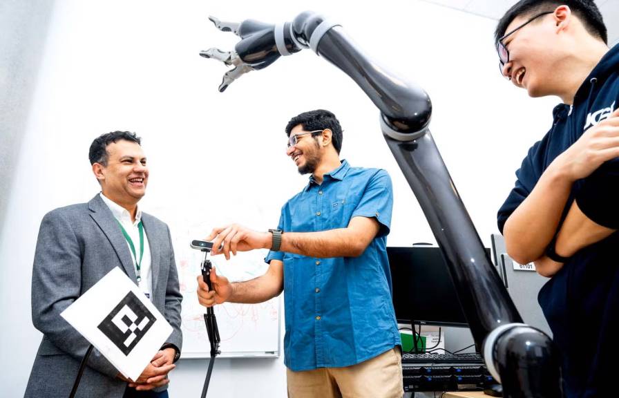 Three people smiling in a lab with a robotic arm in the foreground.