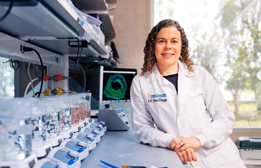 A researcher in a lab coat stands in a laboratory, smiling confidently. Lab equipment are arranged on the counter.