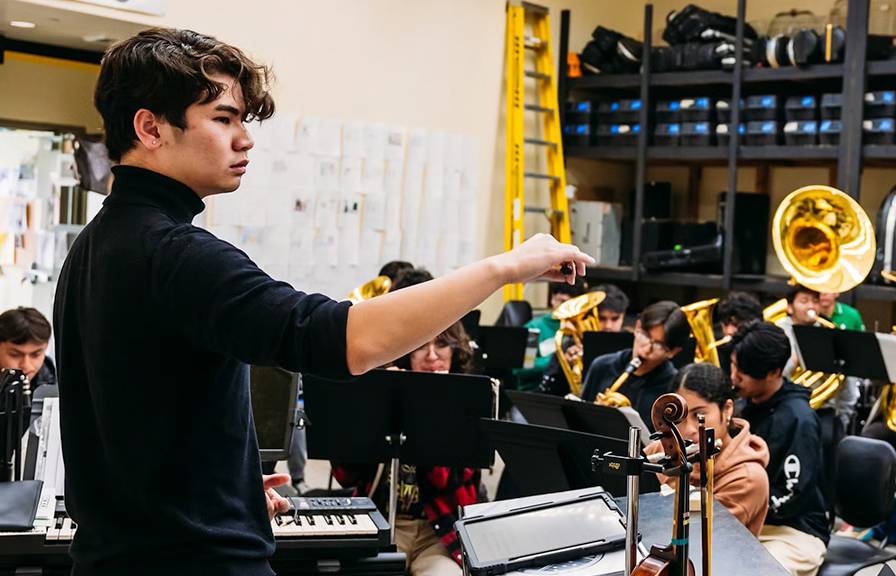 A young man conducts a high school student band in a rehearsal room