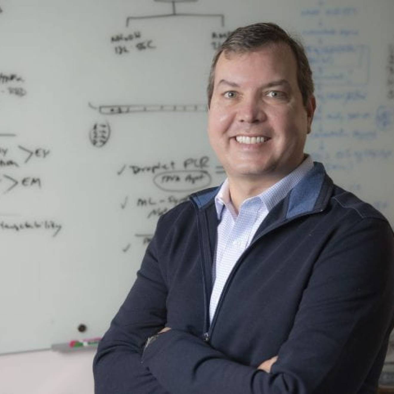 John Chaput, UCI professor of pharmaceutical sciences, stands smiling in front of a white board that is covered with notes.