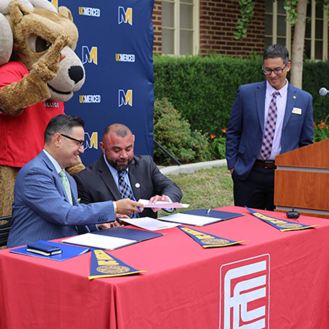 A ram mascot behind two men at a table passing a paper while a man at a lectern looks on