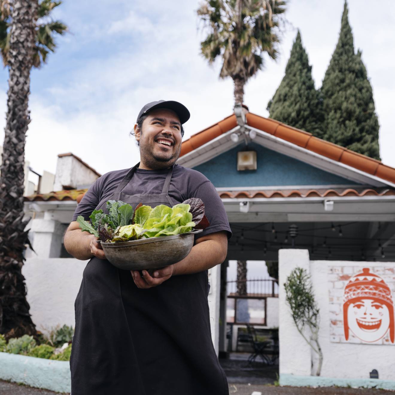 A young man smiles looking up and away from the camera, holding an armful of recently harvested lettuces. He's standing in front of a low green building with palm trees in the background. It looks like LA. 