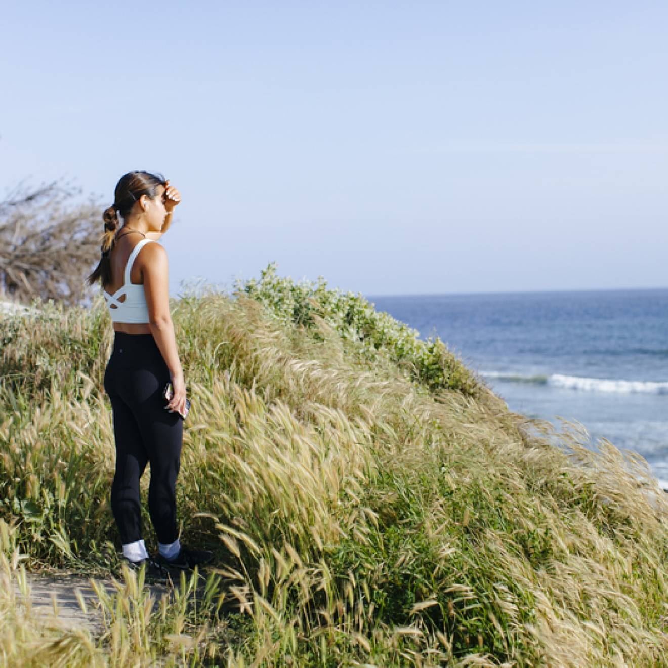 A young woman in exercise attire stands on a grassy bluff overlooking a beach, shading her eyes from the sun