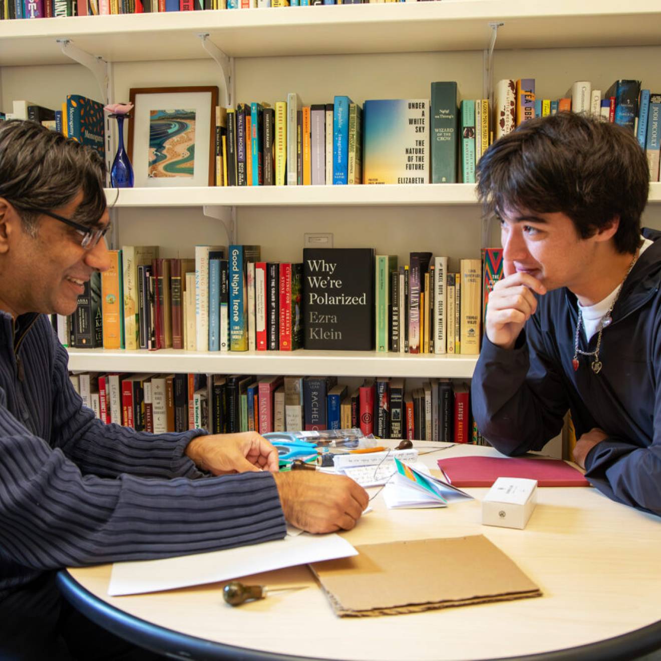 A professor-aged man and a student-aged man sit at a round table in front of a bookshelf, facing each other in discussion. 