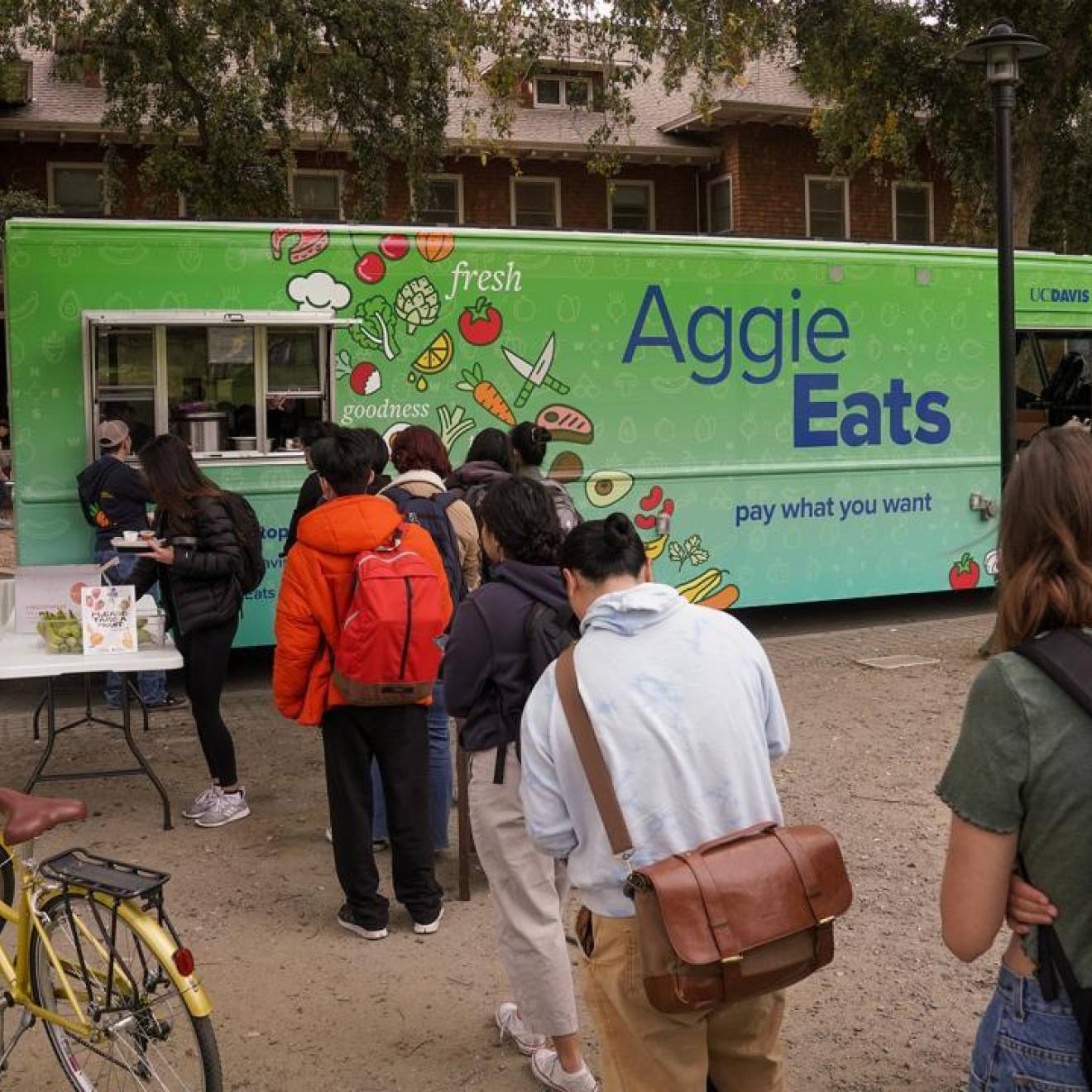 Students with backpacks and bikes line up in front of a food truck. The side of the food truck reads "Aggie Eats. Pay what you want."