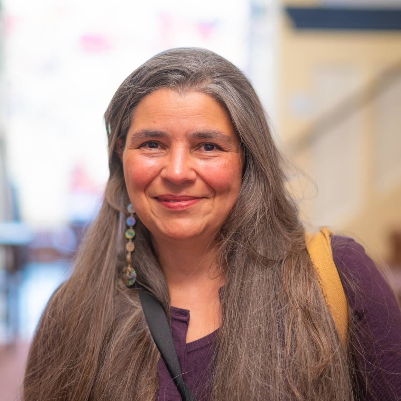 Carolyn Smith smiles at the camera in a shoulders-up portrait with a staircase and window in the background. She has long, straight, light-brown hair tinted with gray and wears a maroon top and long, dangly earrings. 