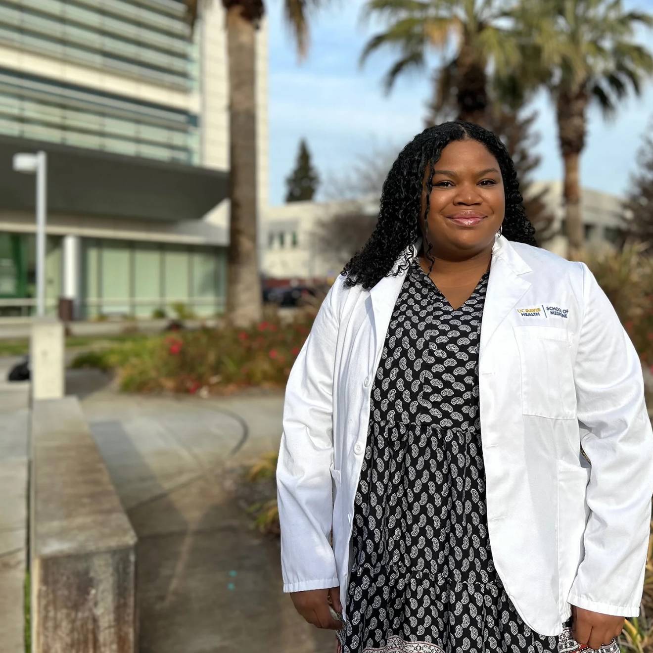 Chelsea Nash wears a black and white polka dot dress and white doctor coat, smiling for a portrait outside of a medical building at UC Davis