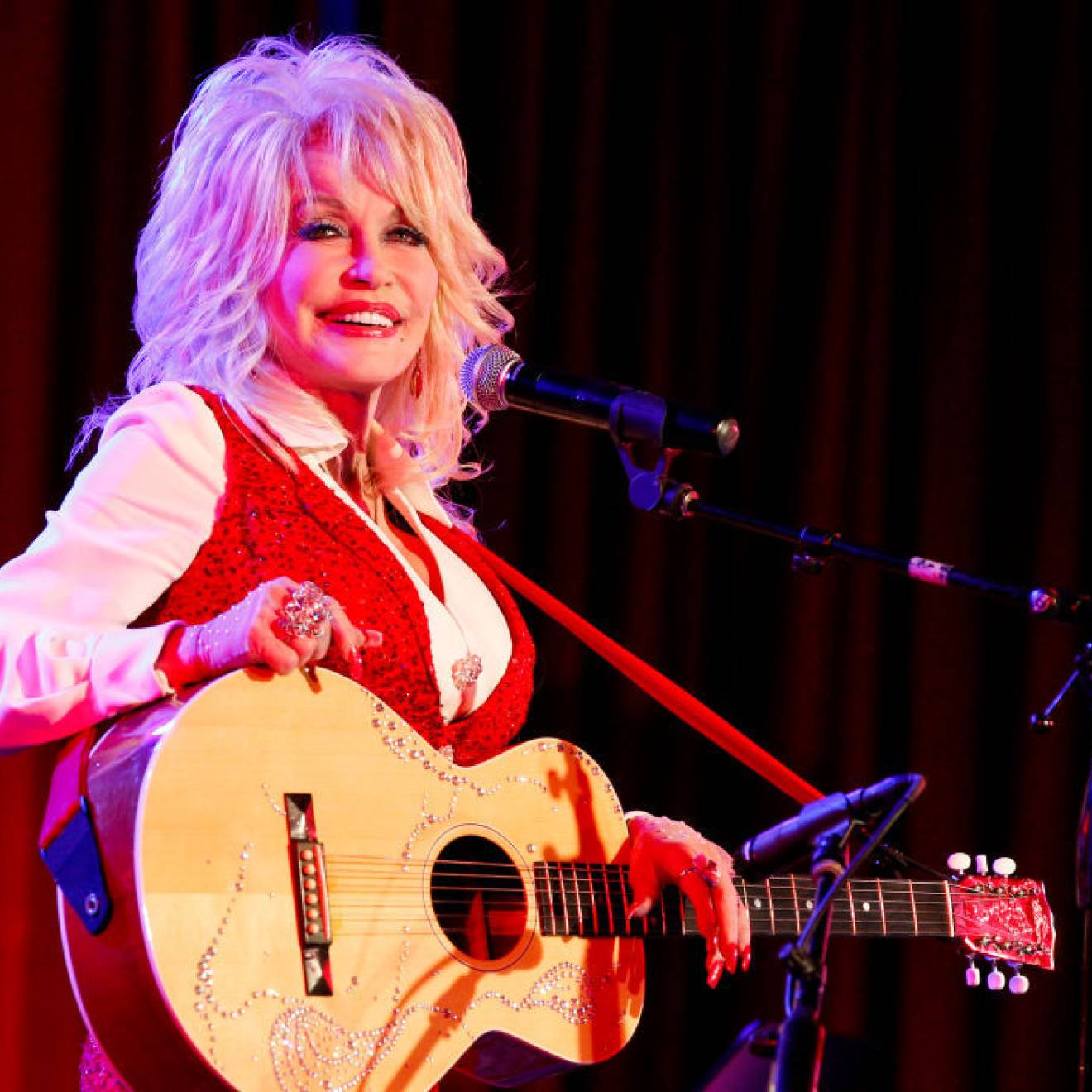 Dolly Parton in a red vest smiles while holding an acoustic guitar