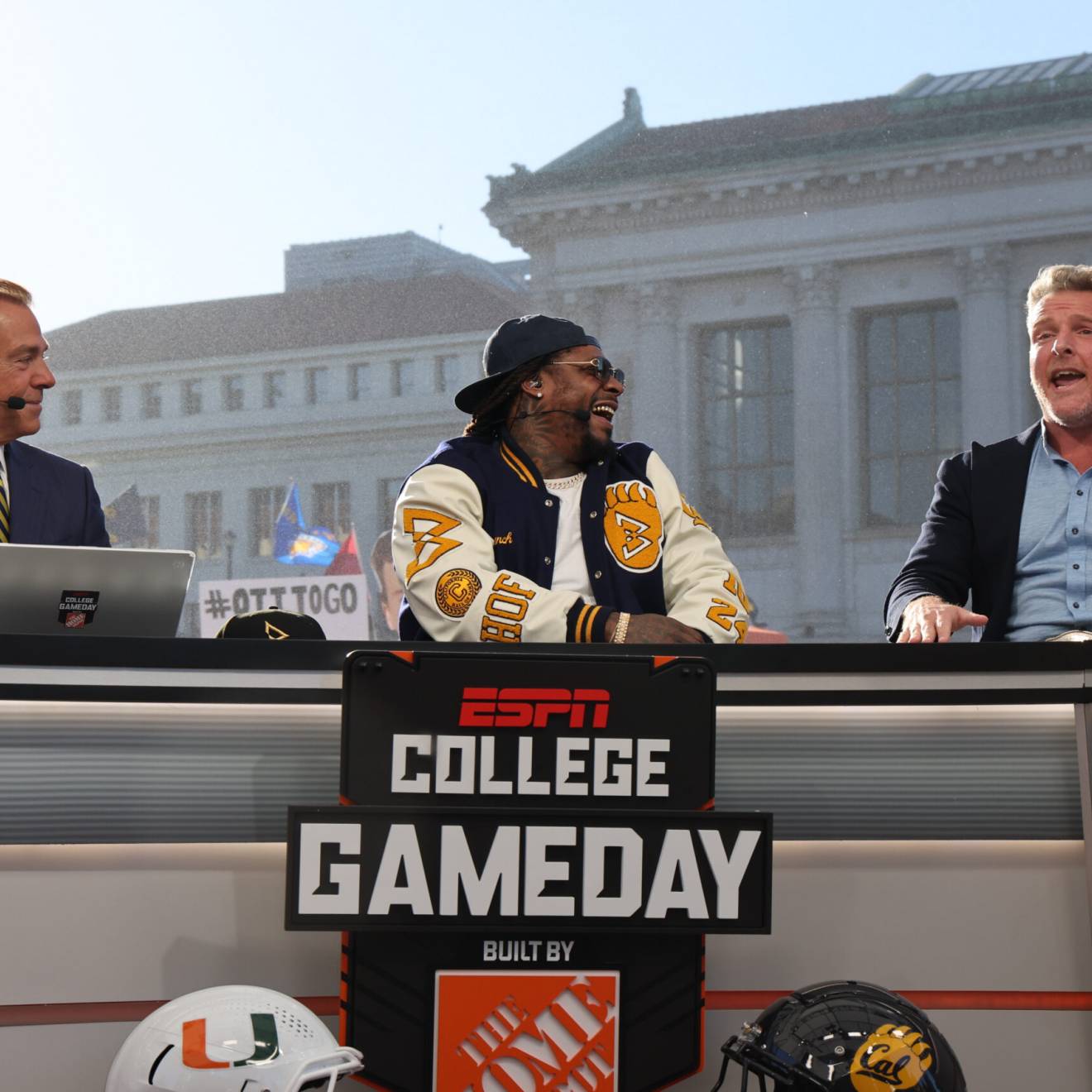 Nick Saban, man in a blue and gold tiem next to Marshawn Lynch, wearing sunglasses, a backwards hat, and a Cal bomber jacket, next to Pat McAfee, man in dress shirt and blazer without tie, mouth open, on the set of College GameDay, on the Cal campus, Campanile and Doe Library visible behind in the morning light