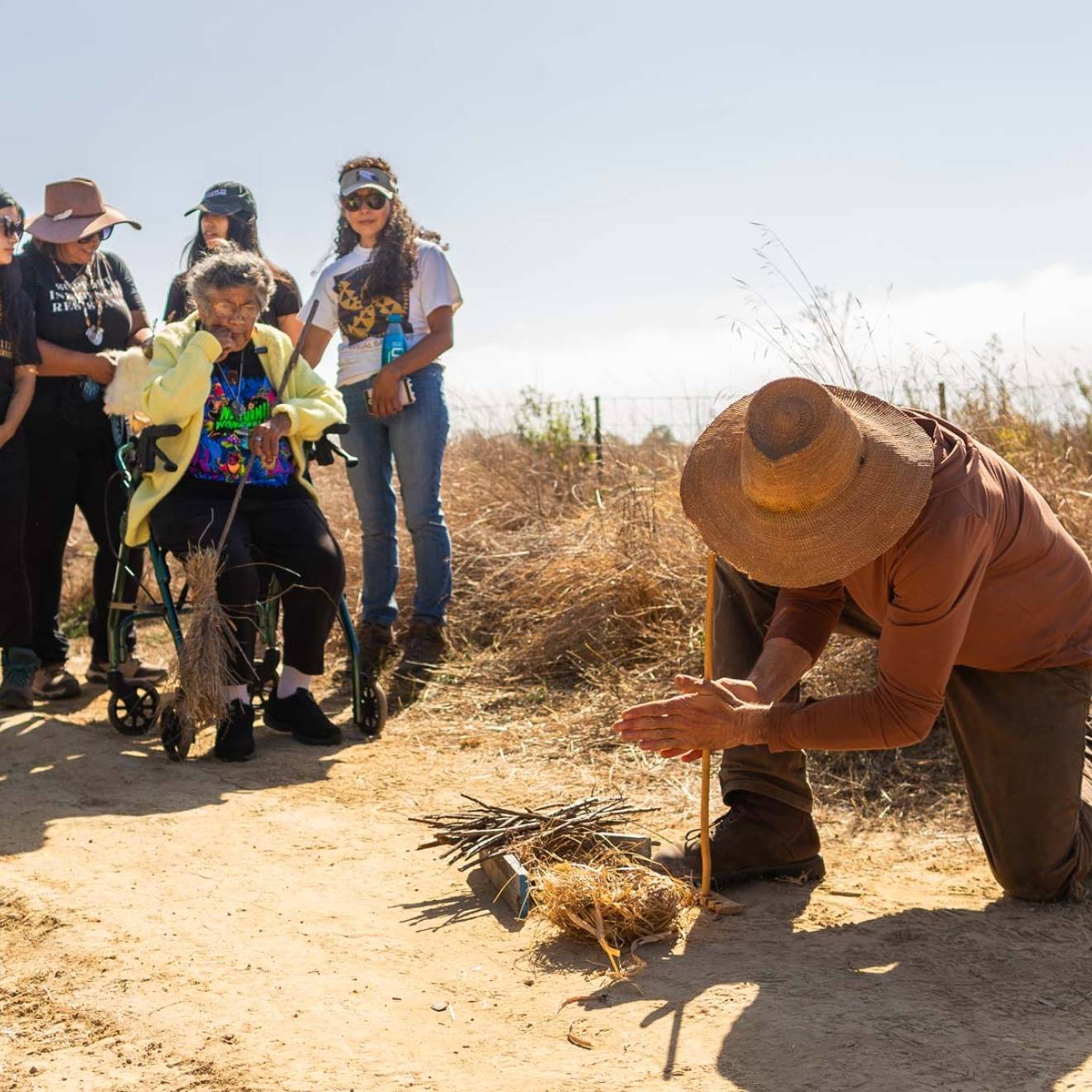 A man in a straw hat uses a wooden stick to start a fire in a brush pile while 6 people watch.