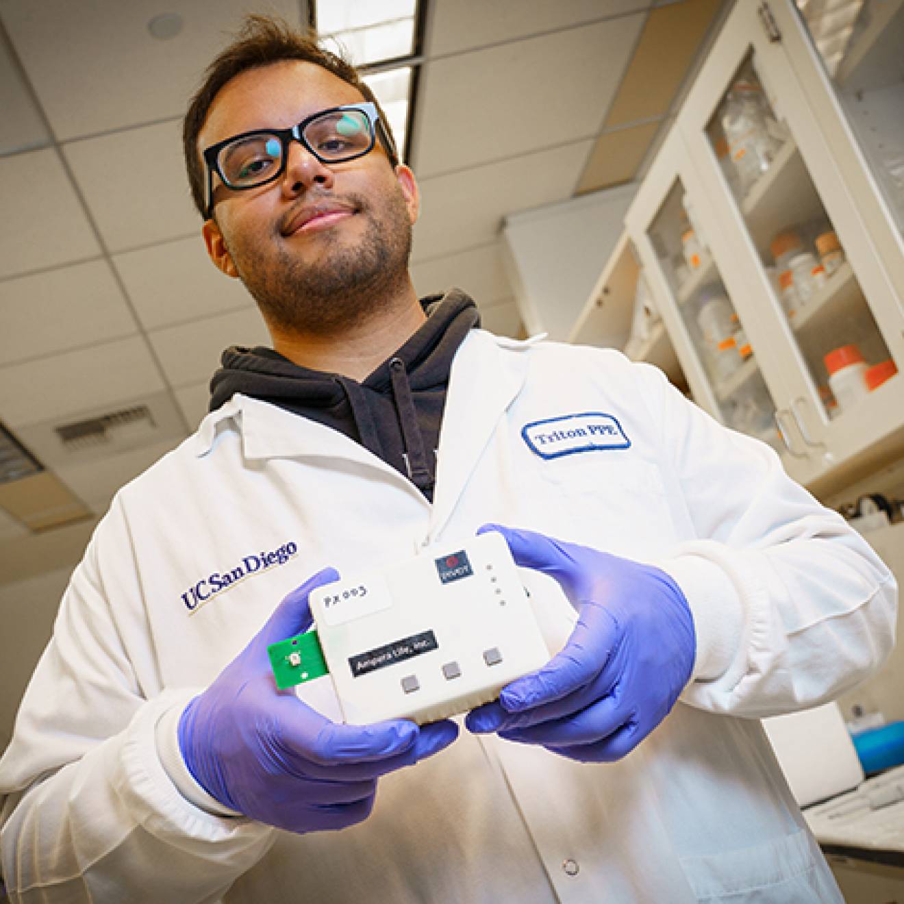 A male student in a white lab coat wearing glasses and purple rubber gloves holds a white box the size of a large sandwich in both hands