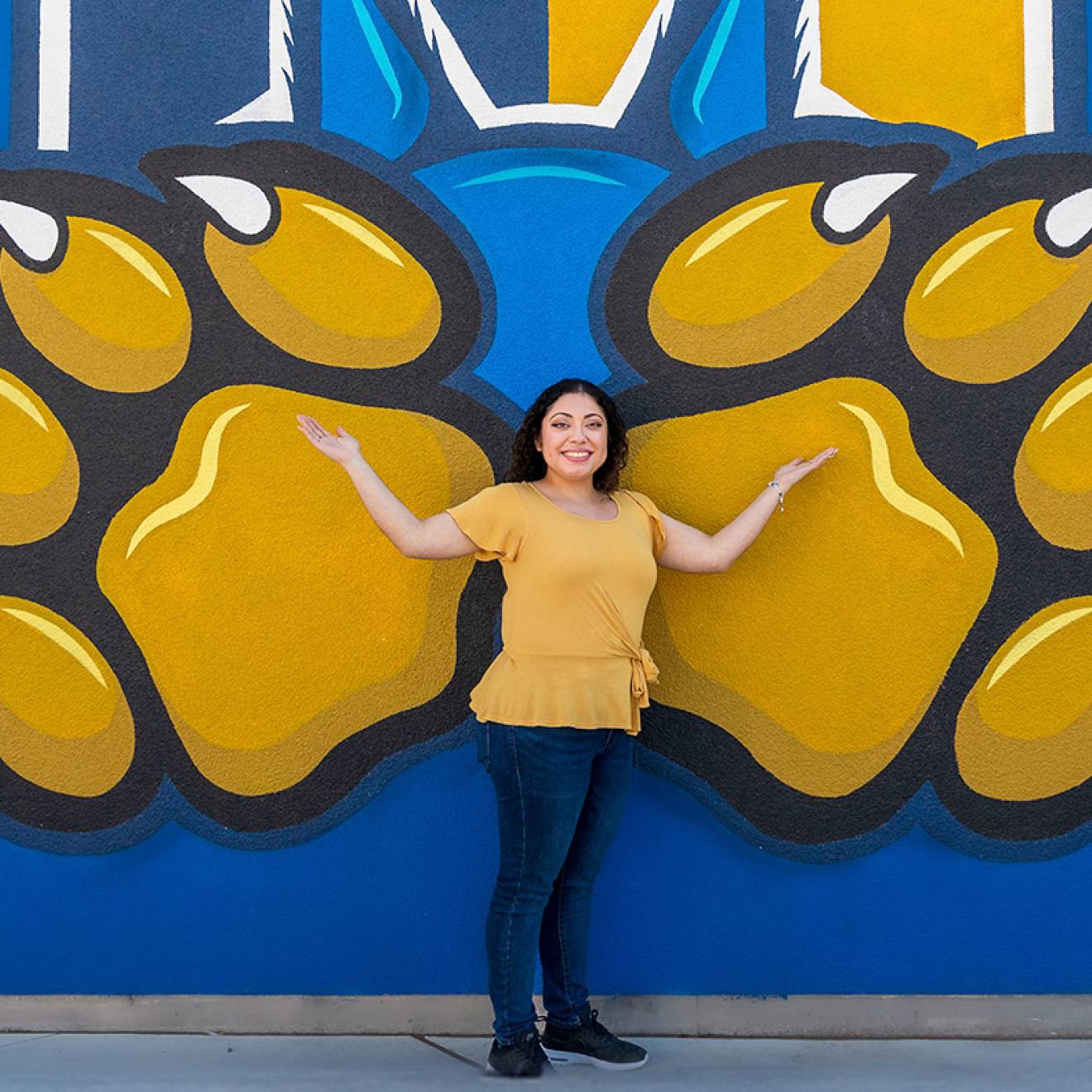 A smiling woman standing in front of a blue wall painted with the UC Merced gold bobcat paws