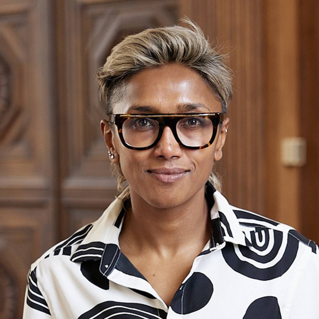 Poulumi Saha, wearing black rimmed glasses and a black and white blouse, smiles faintly for a shoulders-up portrait, with wood paneled walls in the background.