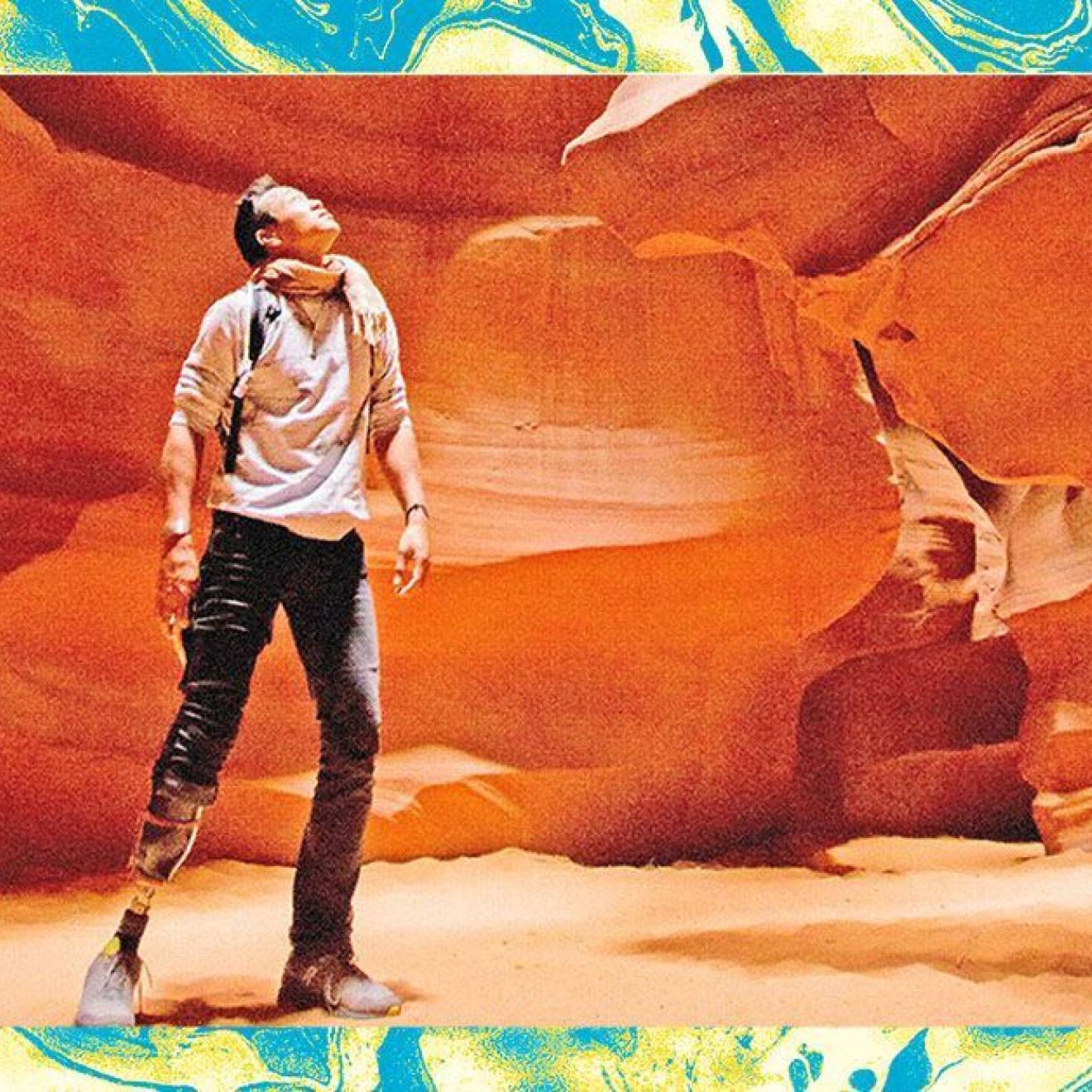 A male hiker with a prosthetic leg below his knee looks up against the backdrop of a red rock canyon wall. The photo is edited and filtered to look grainy, and surrounded by a teal/green textured frame.