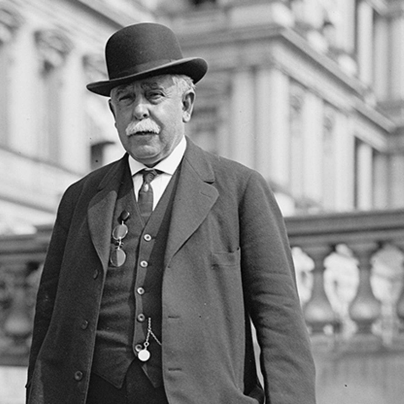 A man with gray hair in a bowler had and three-piece suit in front of a Neoclassical building