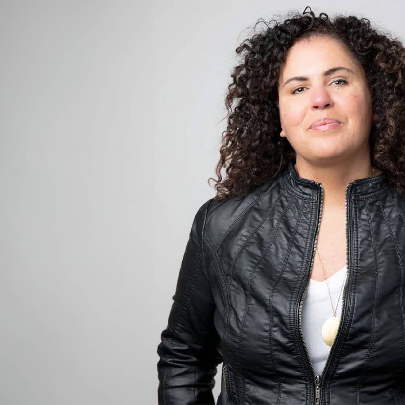 Safiya Noble, wearing a cool black leather jacket, looks resolute in a waist-up portrait against a gray background