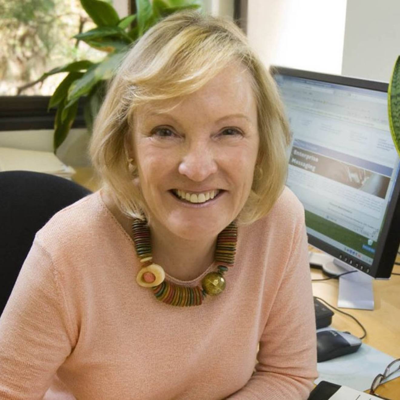 Shelley Taylor, white woman with blonde hair cut in a bob, wearing a pink sweater, smiles at the camera sitting at her desk, with a monitor and a potted planted in the background.