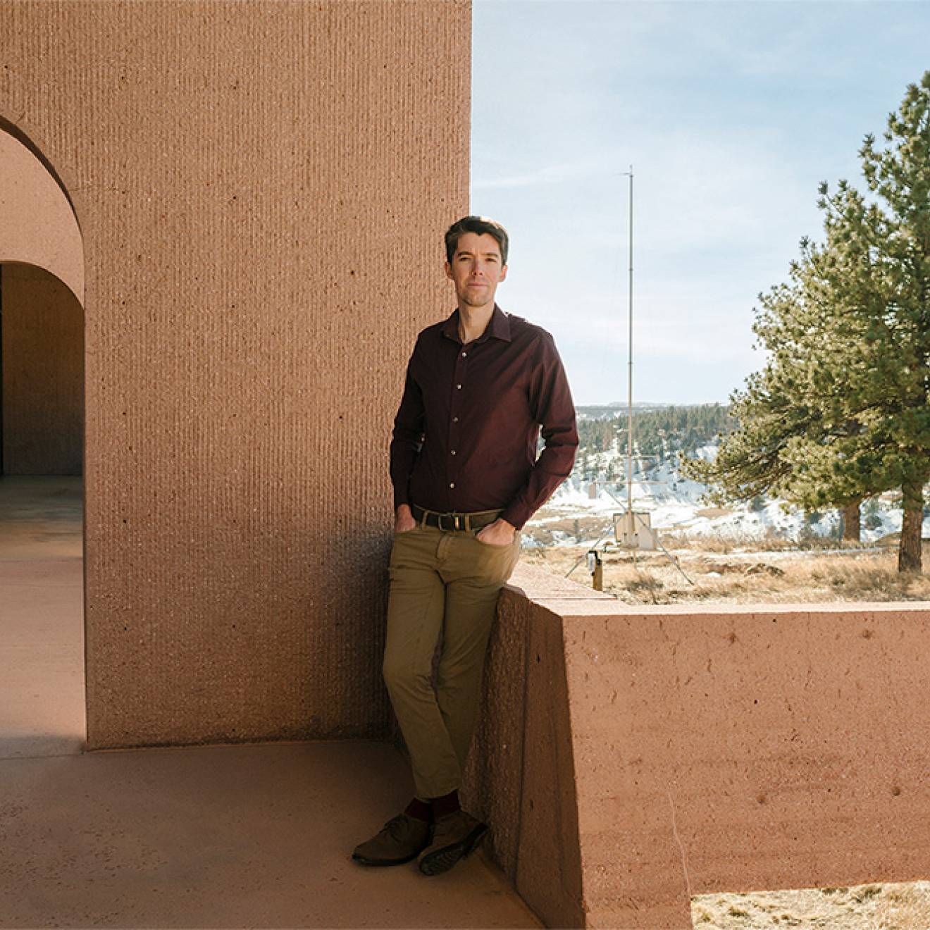 Daniel Swain, wearing khakis and a maroon shirt, leans against a low adobe-colored wall, with a snowy forest in the backdrop and an arched breezeway behind him. 