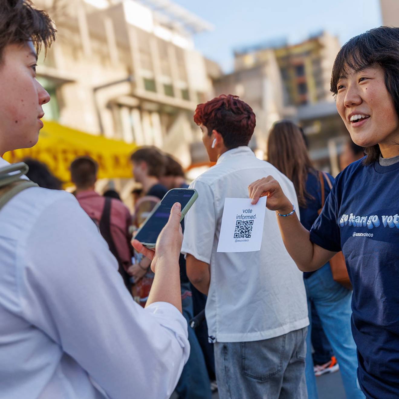 Student wearing a blue shirt reading GO BEARS. GO VOTE. holds a paper with a QR code printed up for a fellow student to scan, on campus at UC Berkeley