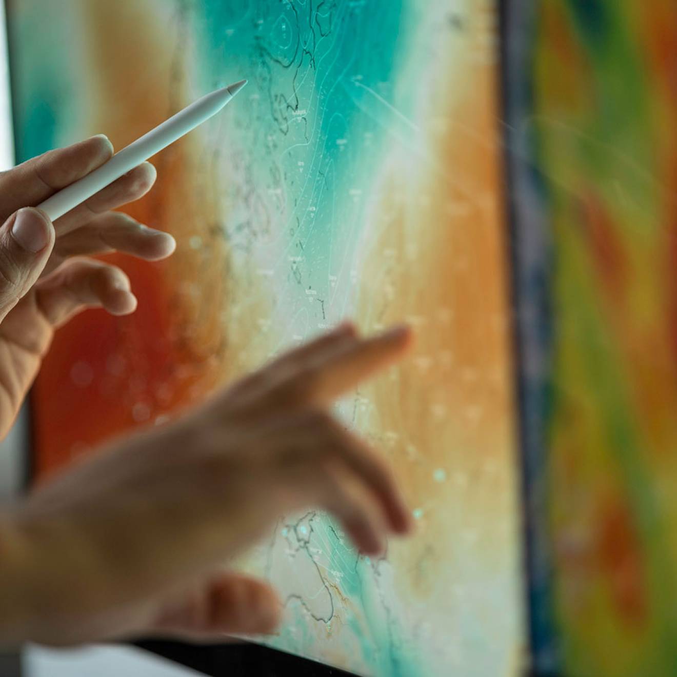 Hands using a stylus and finger to interact with a large touchscreen displaying a colorful weather map
