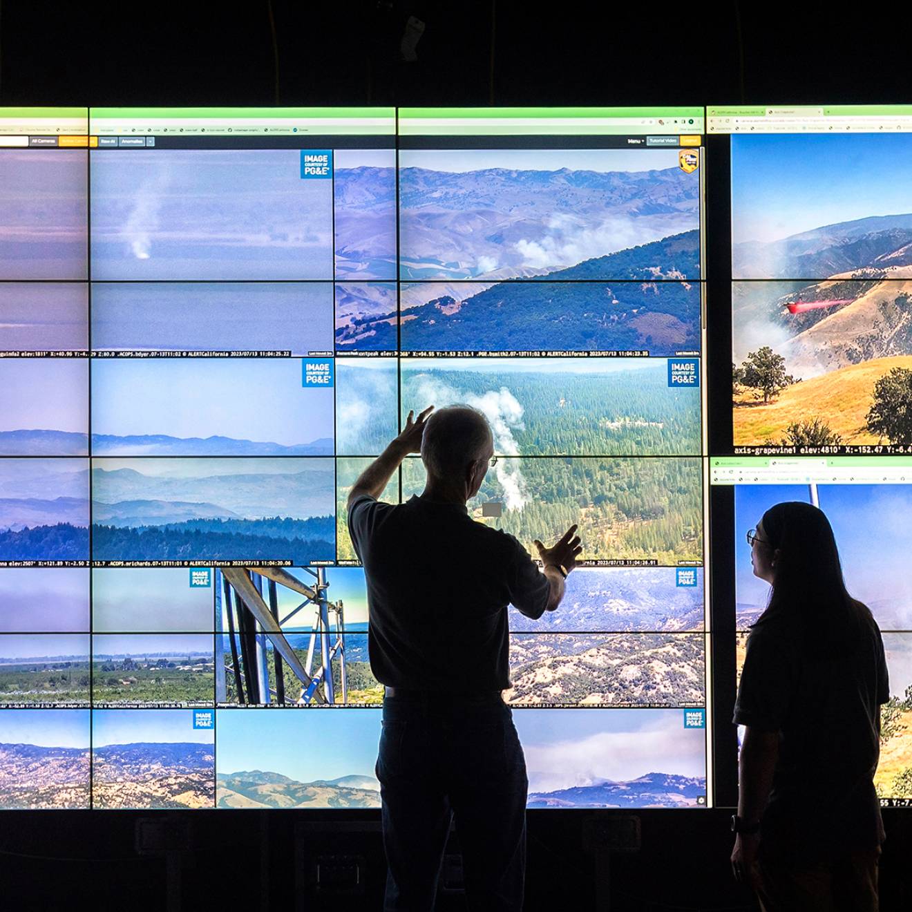 Two people in silhouette look at a large series of screens with visuals of different California landscapes, and a topographical video map of California to the left