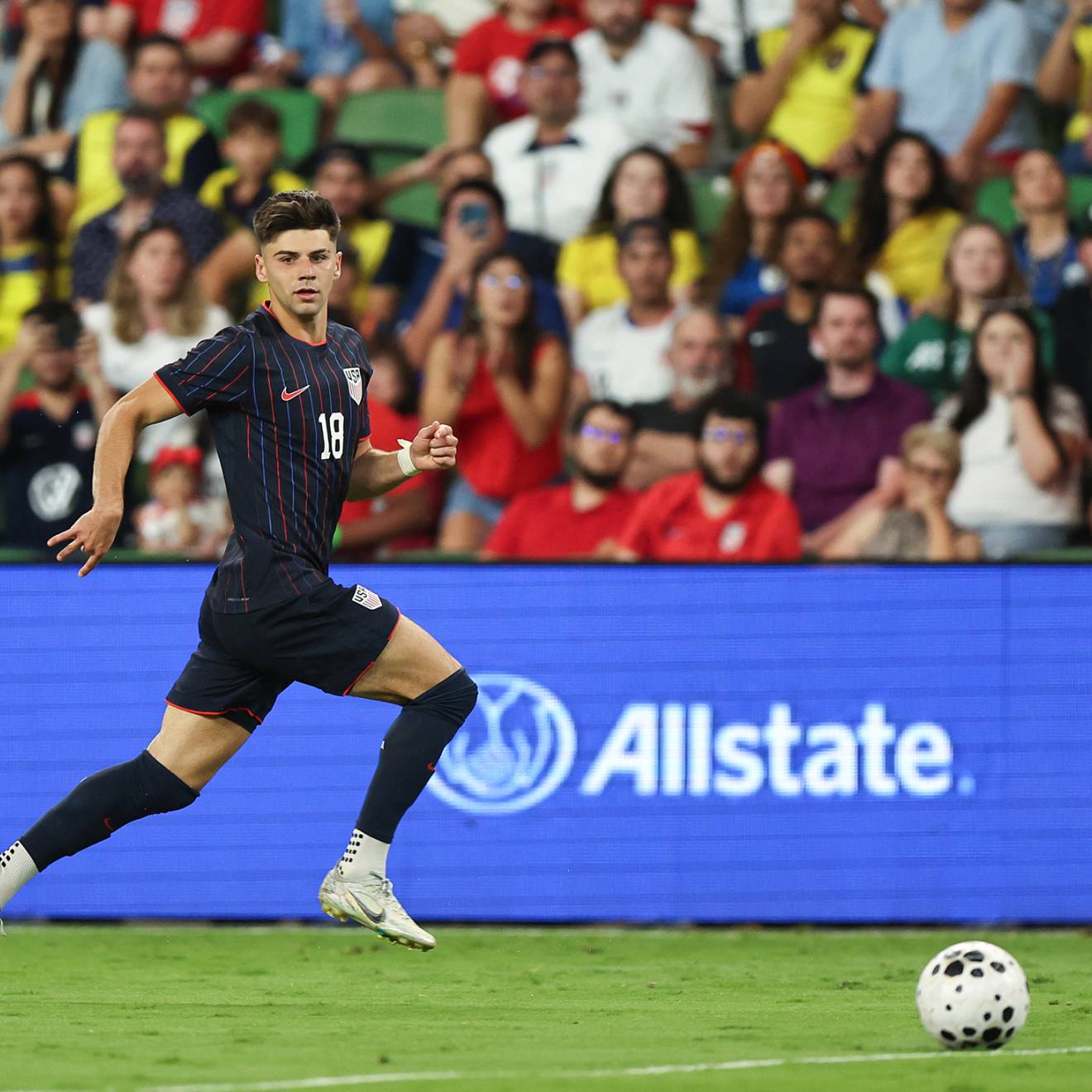 Max Arfsten of the United States in a navy blue uniform with stripes drives a ball during an international friendly match in front of the crowd, looking toward the camera