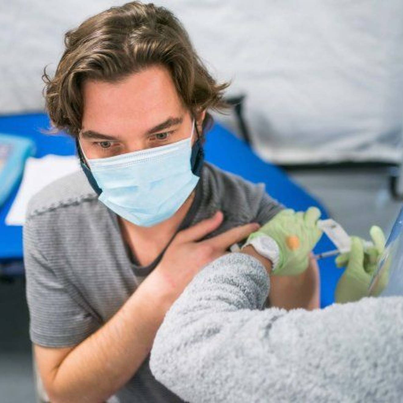 A woman wearing a face shield gives a shot to a young man wearing a face mask