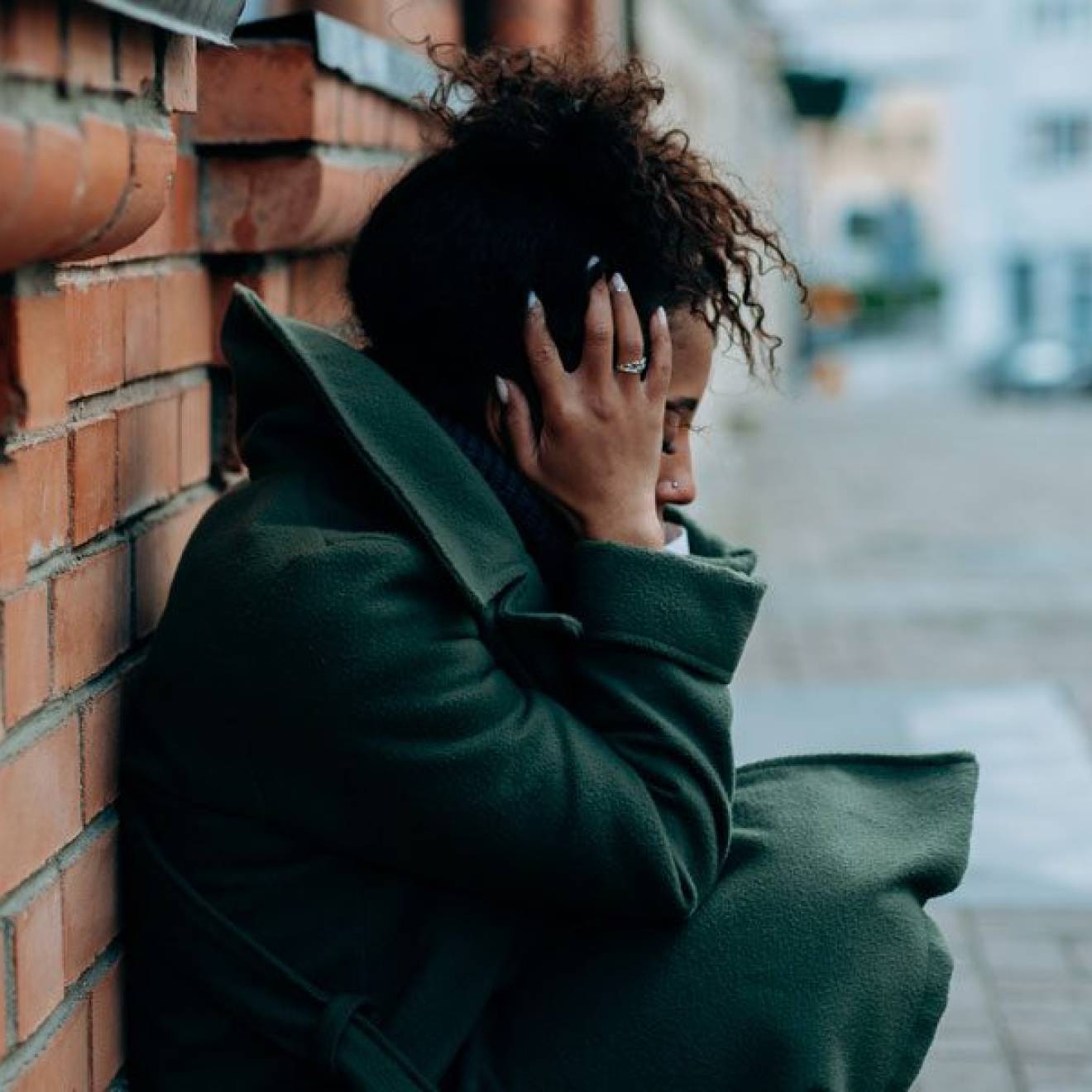 A young Black woman crouches against a brick wall holding her head