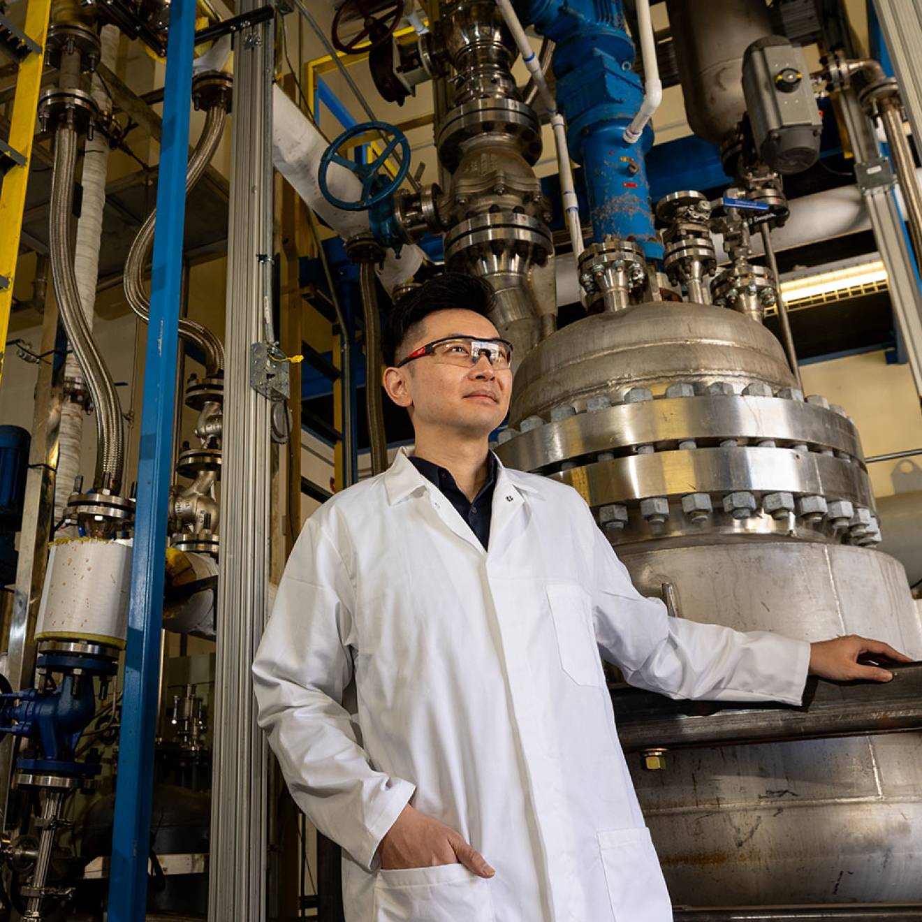 A man wearing a white lab coat and safety glasses stands in an industrial laboratory next to a large stainless steel machine with pipes and bolts, resting one hand on a metal railing.