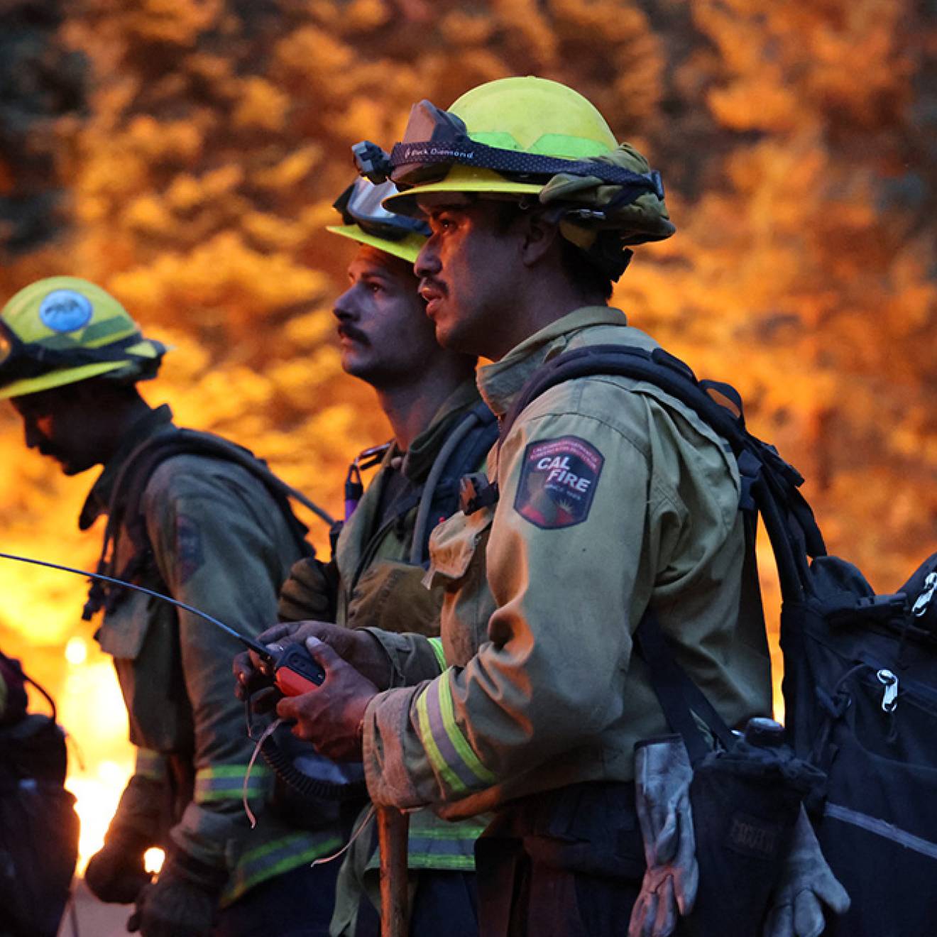 Four firefighters stand against a backdrop of flames. The scene is dark, and 2 young male firefighters hold a device with a long antenna