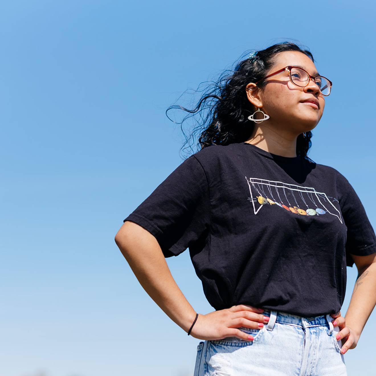 A young woman in a dark t-shirt, jeans and dangly earrings stands with her hands on her hips, looking determinedly forward, shot slightly from below, against a clear blue sky