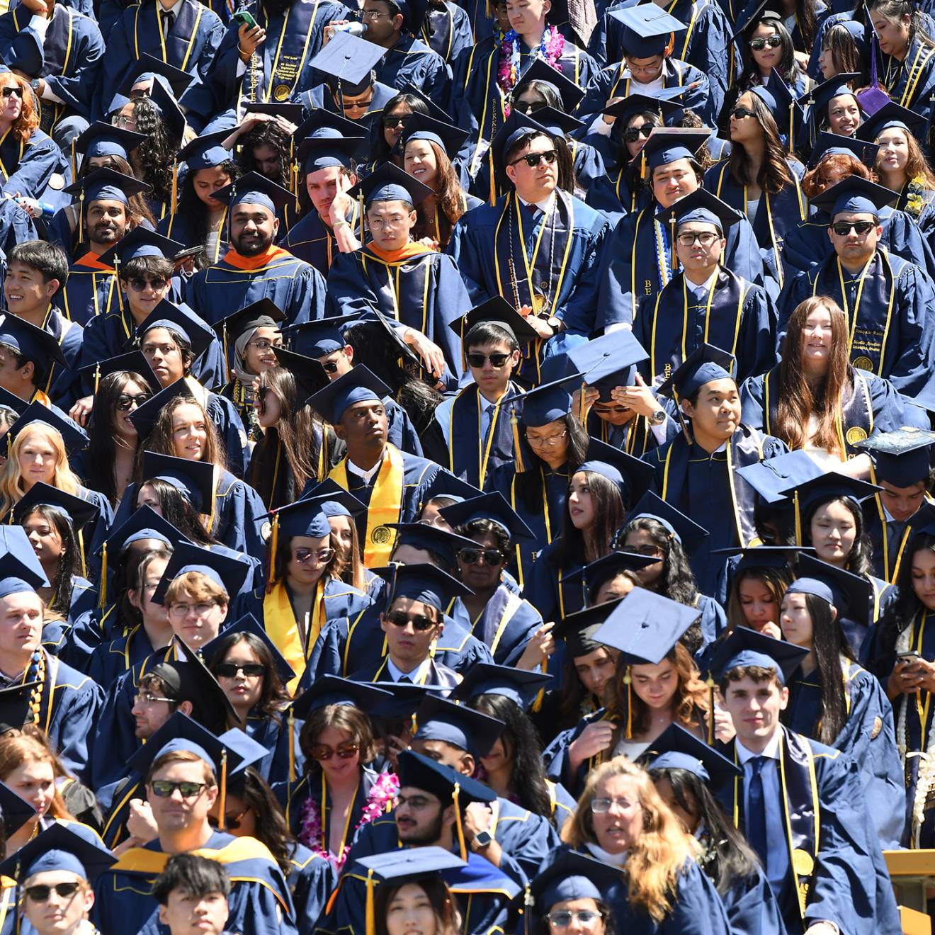 A high-angle shot of a dense crowd of university graduates in blue caps and gowns filling stadium bleachers, accented by colorful leis, stoles and sunglasses under bright sunlight.