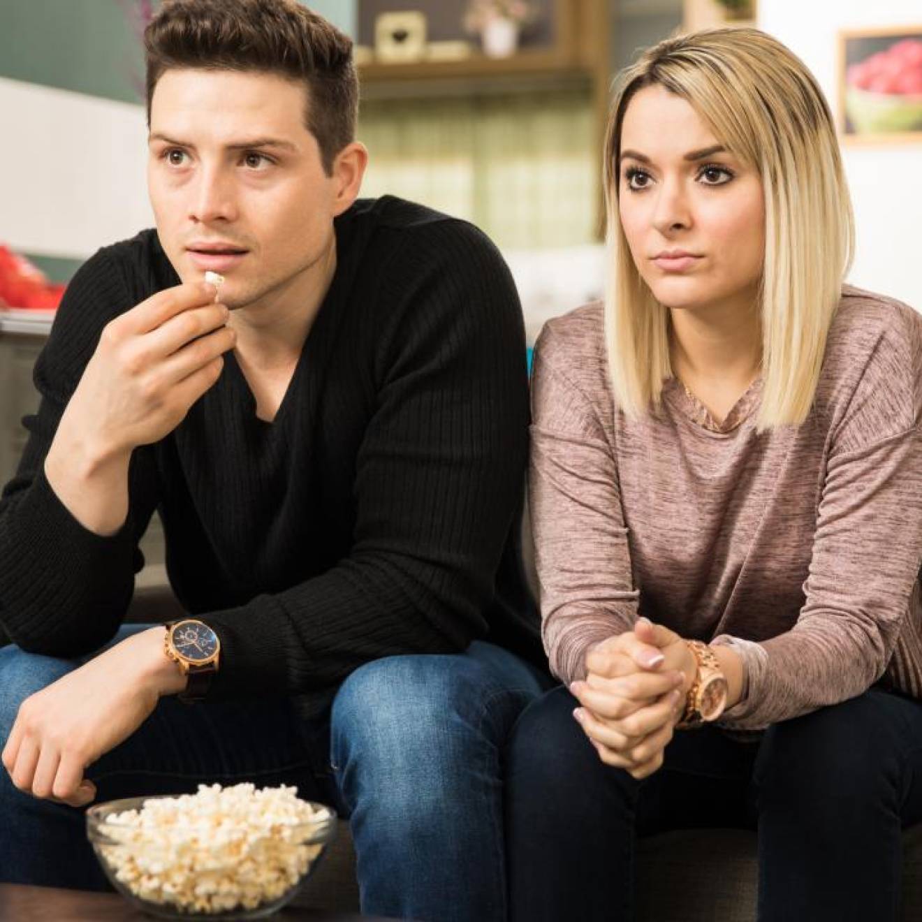 A white man and a white woman watching TV together, looking somber