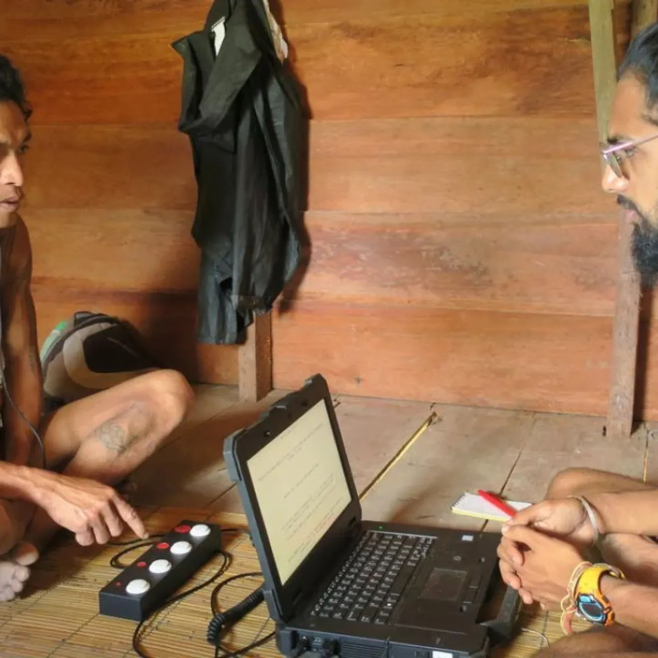 An Indigenous man sits on the floor listening to headphones, facing a researcher with a laptop