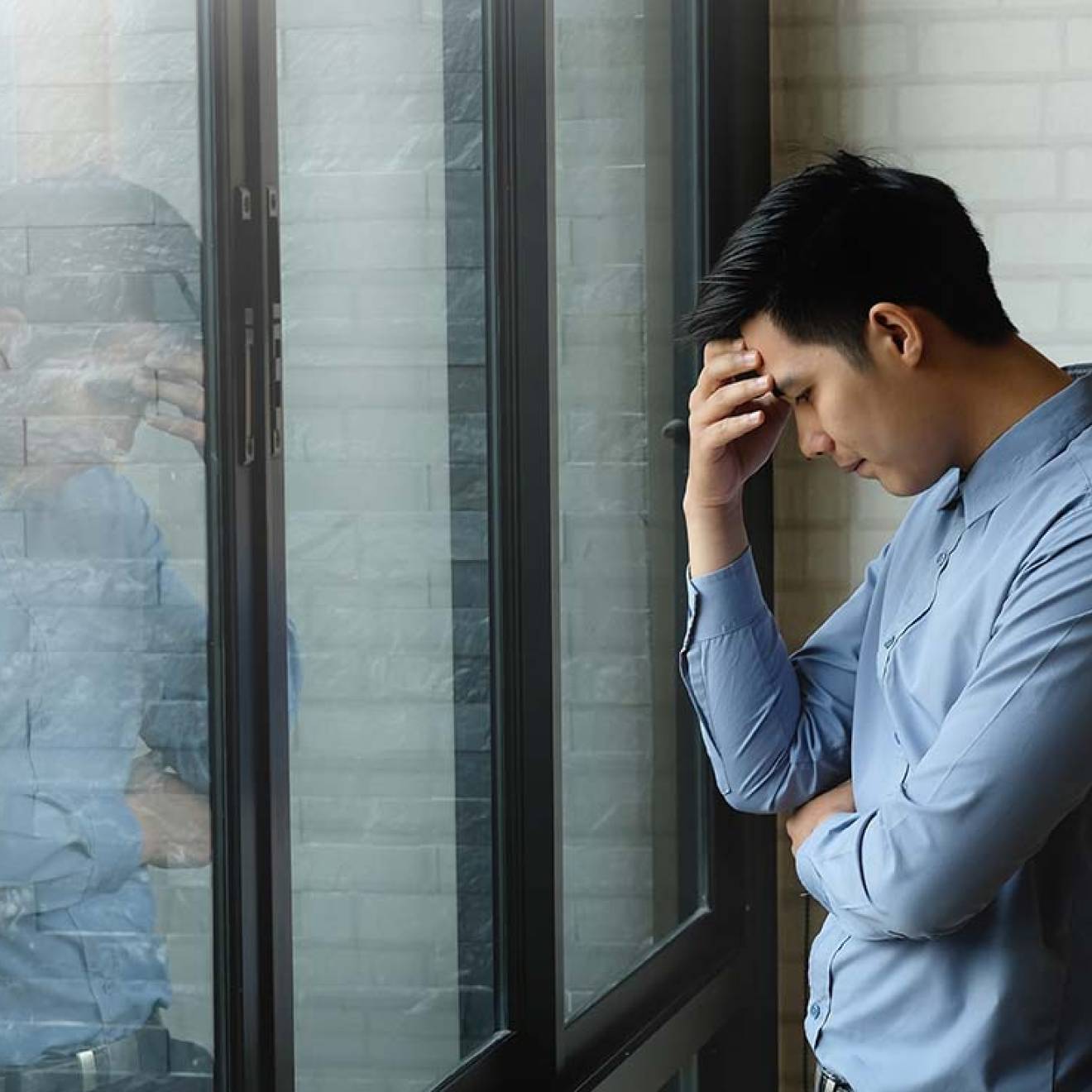 Young Asian man holds head in hand in front of his reflection at work