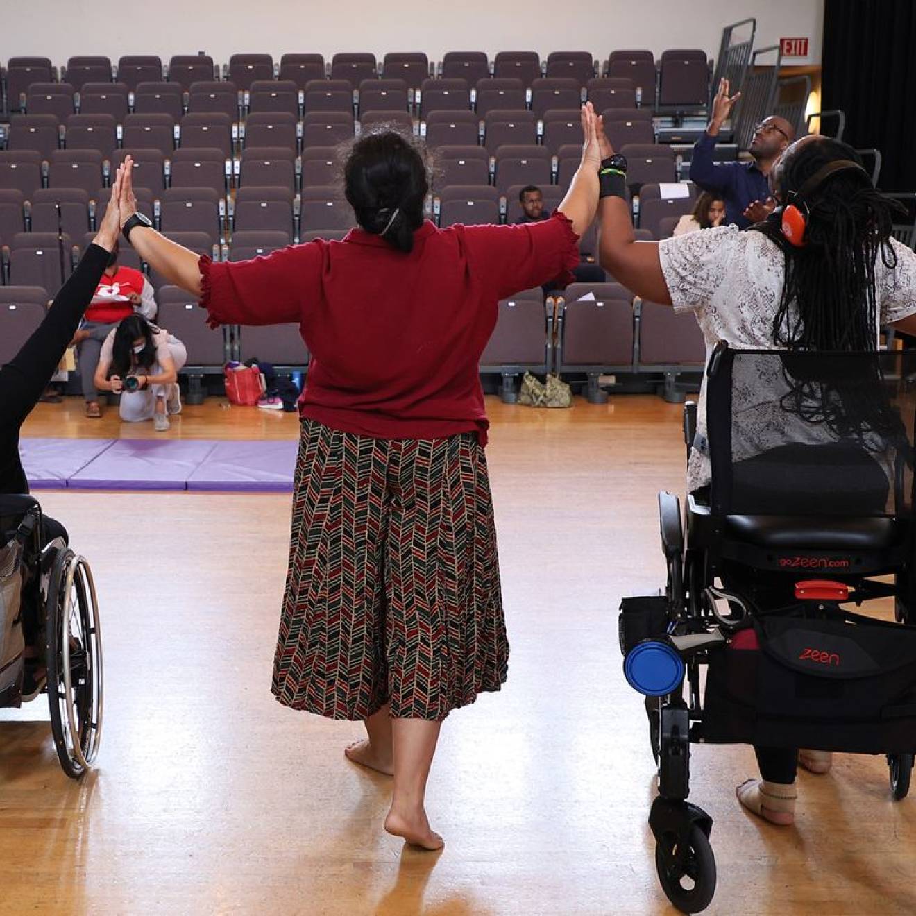 Three disabled dancers, photographed from behind, touch hands while raising their arms.