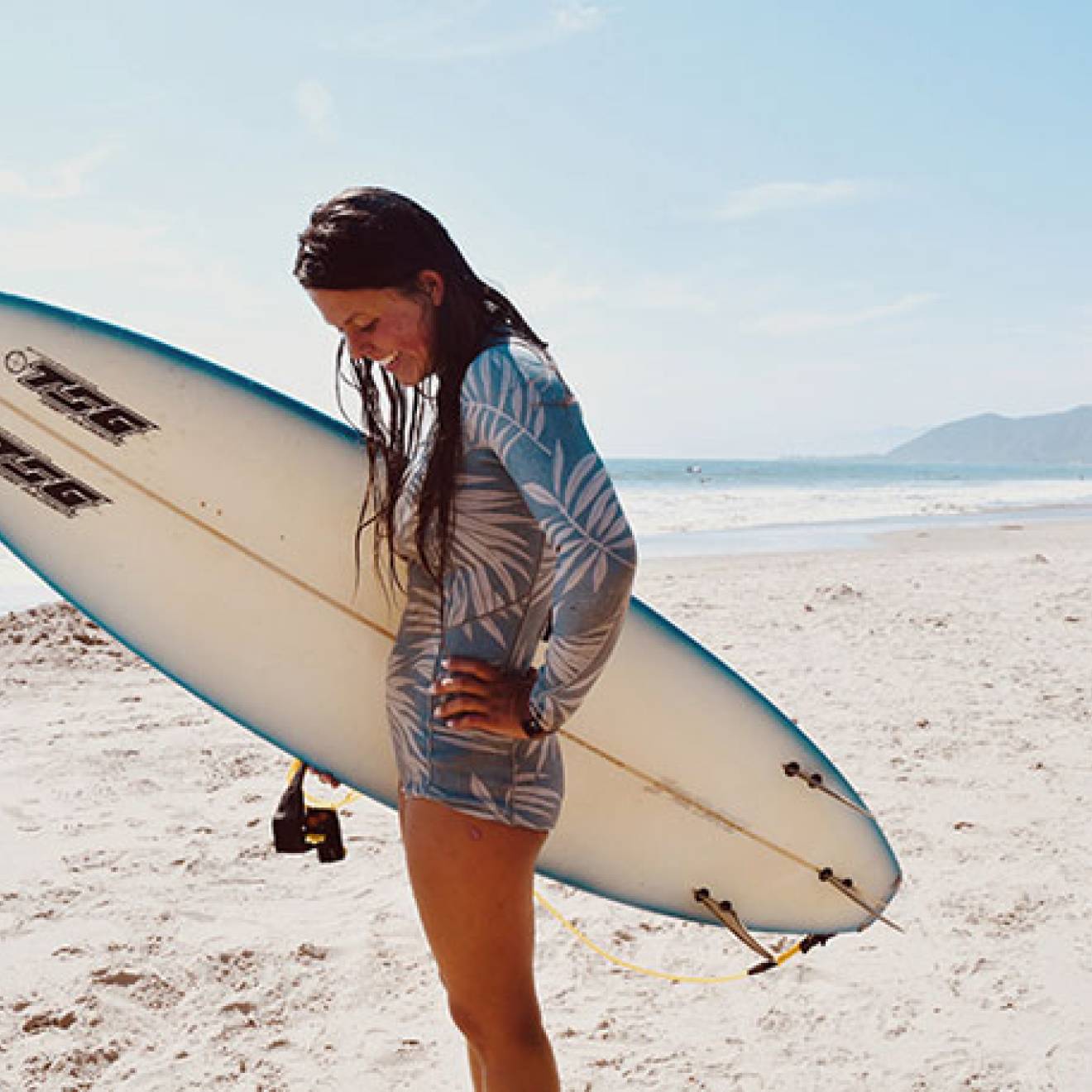 Katia Gibson on the beach with her surfboard looking down, smiling