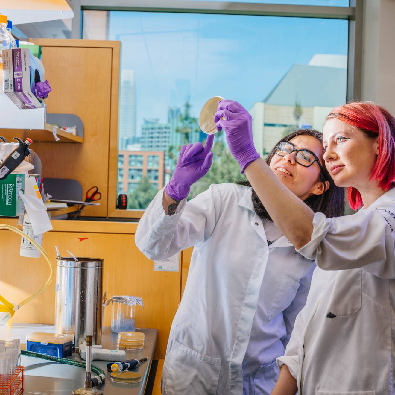 Two female students, one with pink hair, hold up a petri dish in a lab