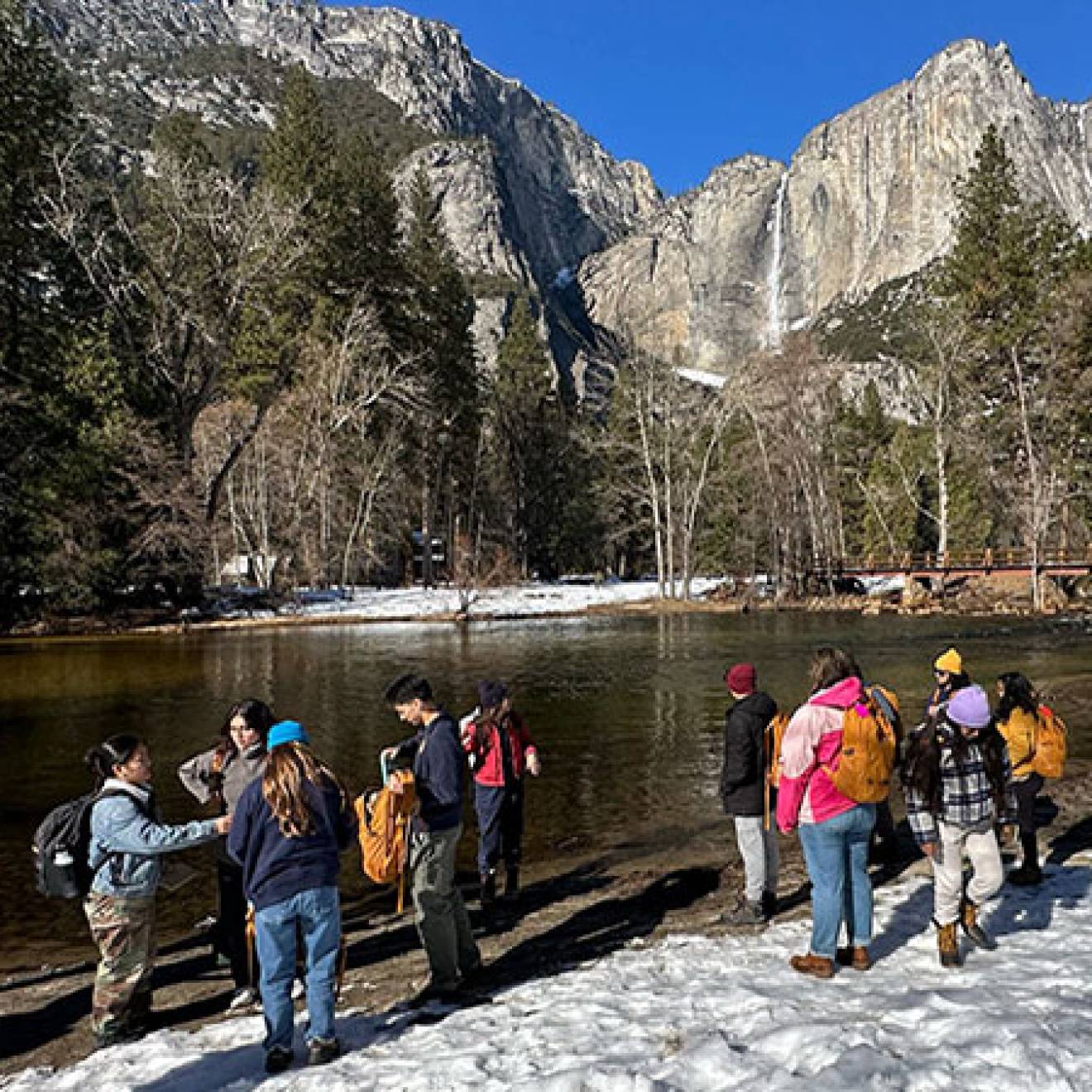 On a sunny day, a group of students gathers on the banks of a river, with snow on the ground and dramatic mountain peaks with a waterfall and forest in the background. 