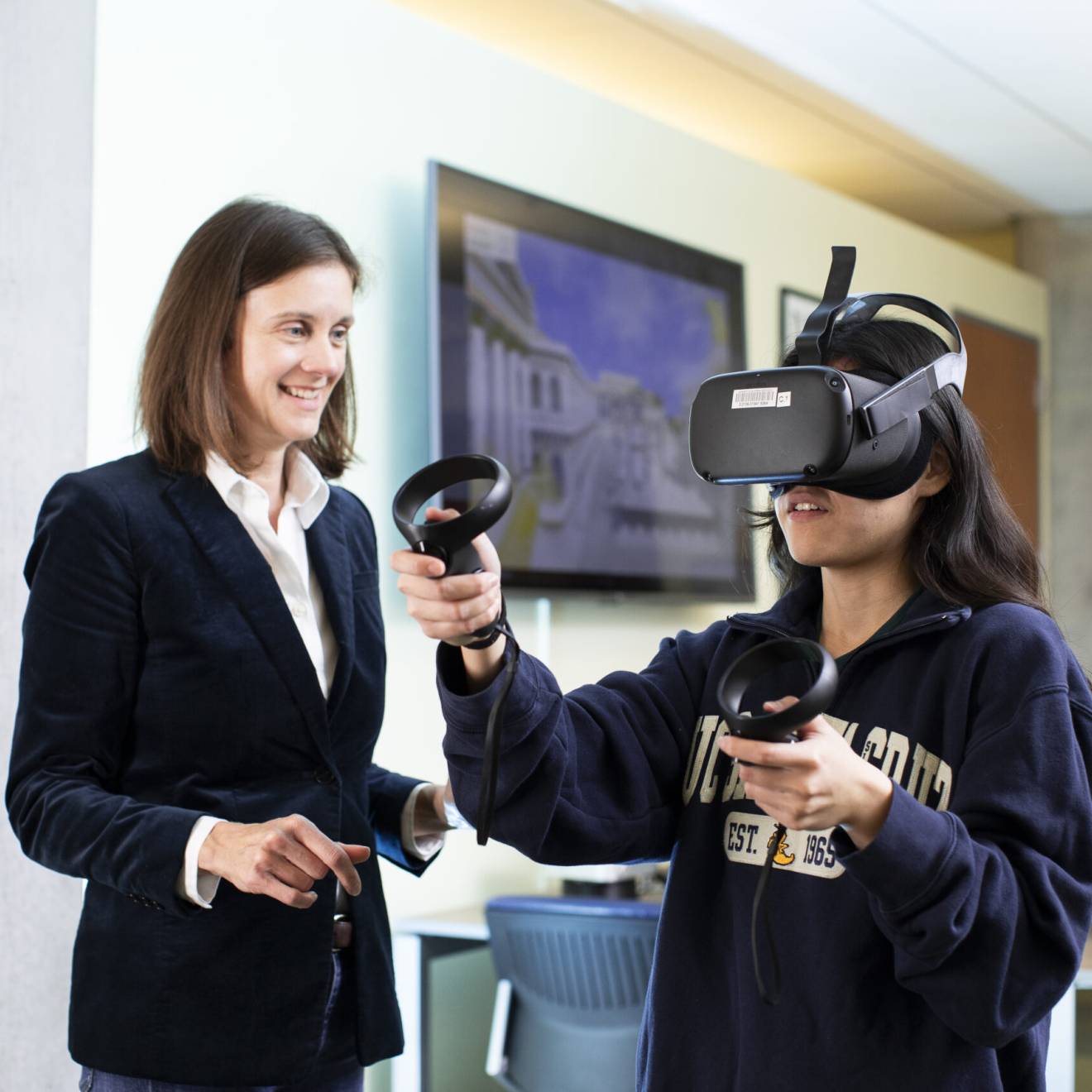 A woman in a jacket and blouse smiles next to someone in a VR headset with a sweatshirt