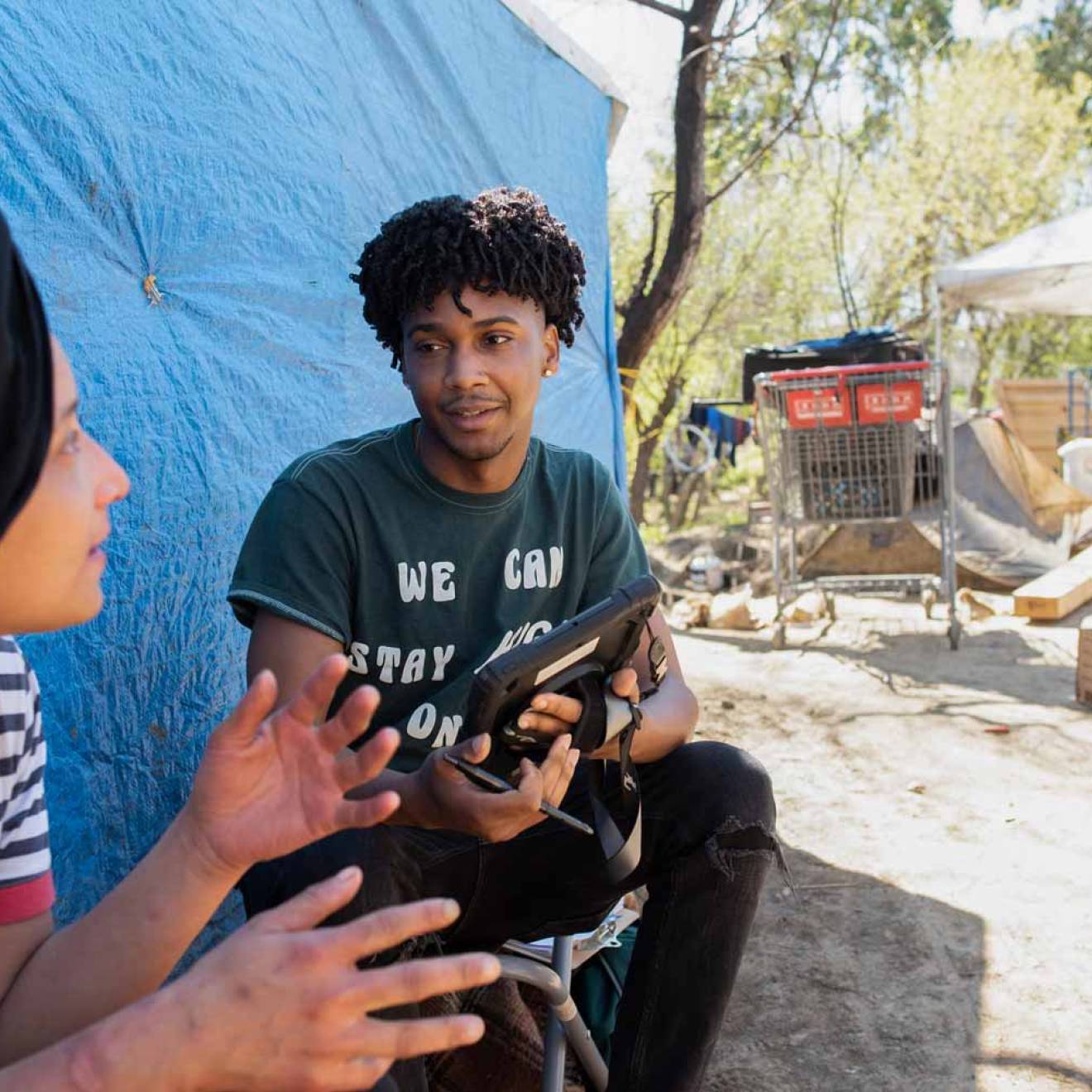 Young, attentive Black man listens to a woman in a homeless encampment and takes notes on a tablet