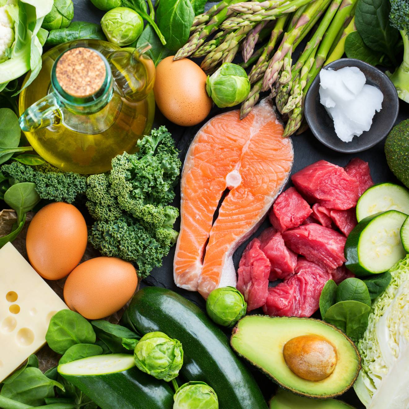 Assortment of healthy ketogenic low carb food ingredients for cooking on a kitchen table. Green vegetables, meat, salmon, cheese, eggs.