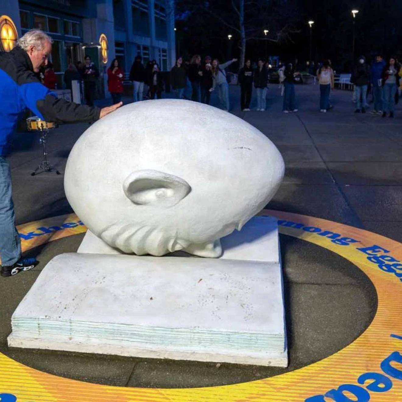 In the front of the photo, a man touches an egghead (a humorous but blank-looking human face shaped like an egg) which is faced down on a concrete open book; in the back of the photo, a line of people outside a museum at night