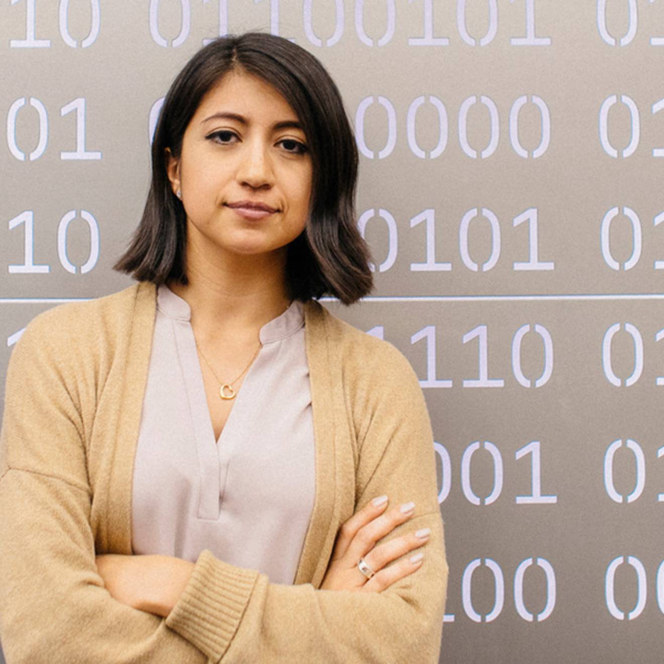 A woman with shoulder length dark hair wearing a cream colored cardigan looks resolutely at the camera with her arms crossed. She's standing in front of a metal panel with sequences of 0s and 1s cut out to simulate computer code. 